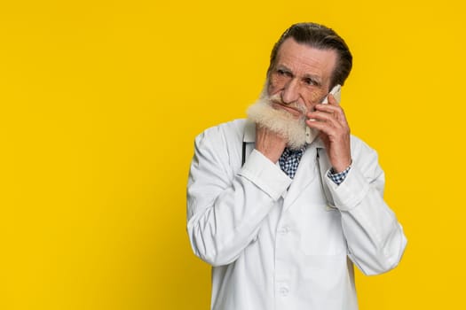 Disinterested senior doctor cardiologist man having annoyed boring talk on smartphone. Tired sleepy apothecary pharmacy grandfather freelancer talking on mobile phone with friend on yellow background