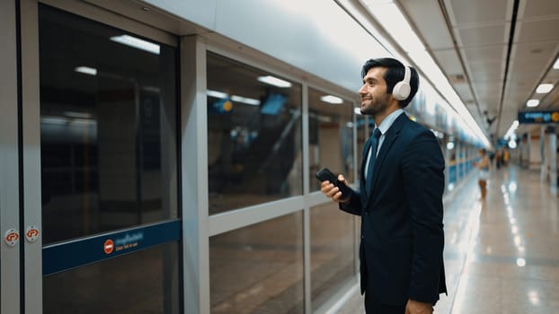 Project manager wearing headphone at train station while holding mobile phone for choosing song. Smart business man listening relaxing music while waiting for train with blurred background. Exultant.