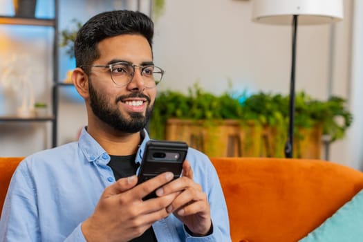Happy young man thinking and text messaging on smartphone. Indian guy sitting on sofa and using cellphone in living room at home. Arabian male is searching information on browser internet. Copy-space
