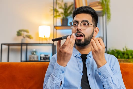 Cheerful Indian man enjoying mobile loudspeaker talking gossip, rumors, good news sitting on sofa. Happy excited Hispanic guy making phone call conversation with friends at modern home room apartment.