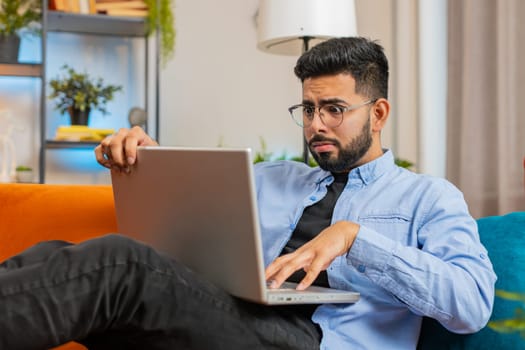Upset young Indian man sitting on sofa at home using laptop having anxiety and stress. Shocked bearded Hispanic guy reacting to loss, bad news on netbook in living room apartment. Computer virus.