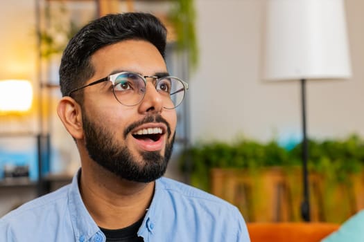 Portrait of amazed shocked Indian man wearing eyeglasses with wow reaction open mouth in living room at home. Happy surprised Arabian guy with big eyes, excited by sudden victory, good win news.