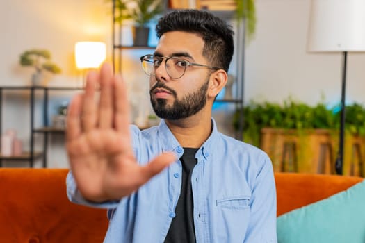 Portrait of Hispanic young man guy on sofa with serious expression doing stop gesture with hand at home room, warning of finish, prohibited access, declining communication and body language danger