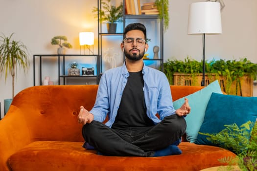 Positive peaceful Indian man taking wireless earphones out of box, placing gadgets in ears, starting media player, listening to music, closing eyes, enjoying leisure, relaxation, meditation at home.
