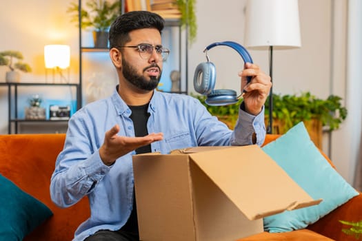 Dissatisfied annoyed Indian male customer open cardboard box receive damaged broken wireless headphones, shocked young man consumer having problem complaint frustrated with bad order delivery at home.