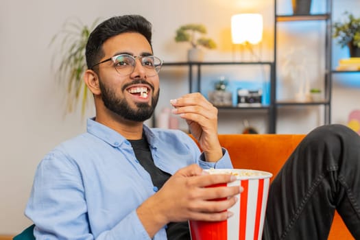 Relaxed Indian young man eating popcorn and watching movie wow expression sitting on sofa in living room at home. Happy Arabian guy in casual clothes enjoying film during weekend in apartment room.
