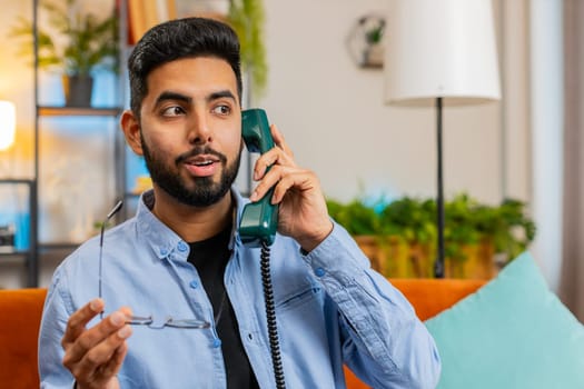 Portrait of shocked Indian man making telephone conversation with friends sitting on comfortable couch at home. Happy excited Hispanic guy enjoying old-fashioned retro phone from 90s talking indoors