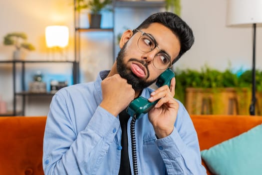 Irritated young Indian man talking on vintage retro wired telephone at home apartment. Disinterested Hispanic guy having annoyed boring conversation with hotline helpline service sitting on sofa.