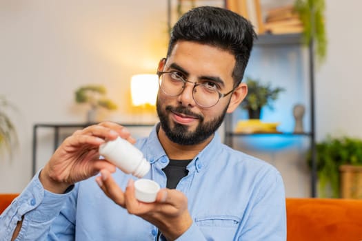 Indian young bearded man taking medicine pill with water sitting on cozy couch at home. Hispanic guy takes medication prescribed by his physician. Medicine, health care, pharmacy and people concept