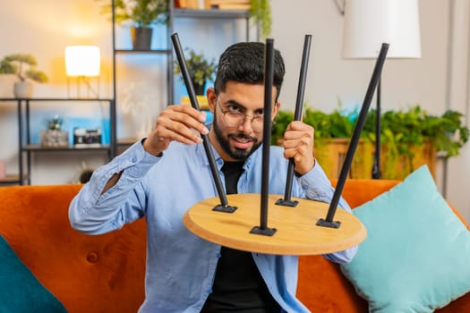 Happy Indian man finishing assembling furniture at home. Successful table desk collect, repair fixing. Hispanic guy after moving into new apartment. Advertisement of a furniture store. Mortgage.