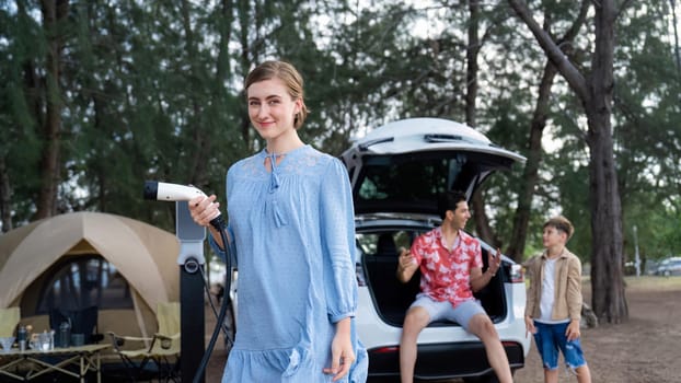 Outdoor adventure and family vacation camping at sea travel by eco friendly car. Cheerful woman or mother holding, pointing EV charger point with playful and happiness posture in campsite. Perpetual