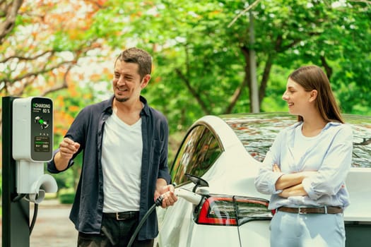 Lovely young couple recharging battery for electric car during autumnal road trip travel EV car in autumnal forest. Eco friendly travel on vacation during autumn. Exalt