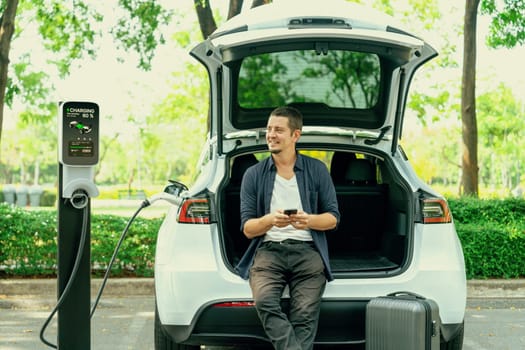 Man using smartphone online banking application to pay for electric car battery charging from EV charging station during vacation holiday road trip at national park or summer forest. Exalt