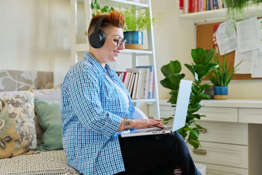 Middle aged woman freelancer in headphones using laptop computer sitting on couch at home. 40s female looking at screen video chat conference call online meeting training mentoring technology for work