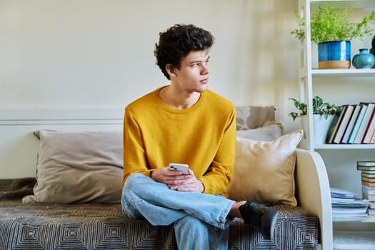 Young guy holding smartphone in hands sitting on couch at home. Handsome university college student 19-20 years old in yellow with curly hair. Technologies, mobile apps, study communication leisure