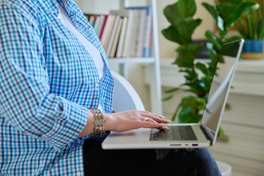 Close-up of mature woman's hands typing on laptop, home interior background.