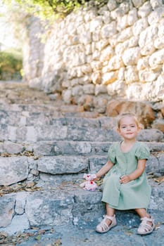 Little smiling girl with toy pink mouse sitting on old stone steps in the garden. High quality photo