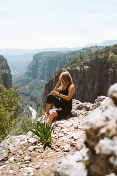 Young beautiful girl woman in black sporty slim outfit sitting on edge rock mountain cliff peak with view on valley canyon. Picture of fit female hiker reached the mountain top.