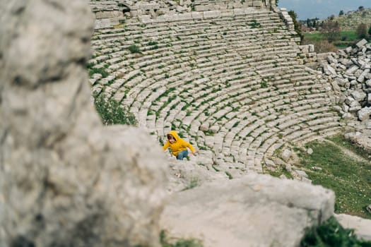 School boy kid in yellow hoodie and blue jeans exploring remains of ancient arena stone coliseum amphitheater.