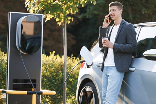 Charging electric car. Portrait of handsome joyful 25-aged business man which has mobile conversation while luxurious electric car charging battery on specially equipped charging station.