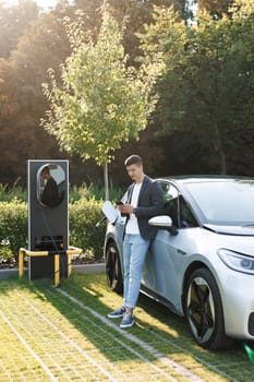 Plug charging an Electric car from charging station. Charging technology. Caucasian businessman using smart phone and waiting power supply connect to electric vehicles for charging the battery in car.