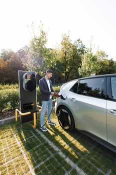 Man is unplugging in power cord to an electric car at sunset. Man charging electric car at charging station using smart phone app. Male unplugging electric car from charging station.