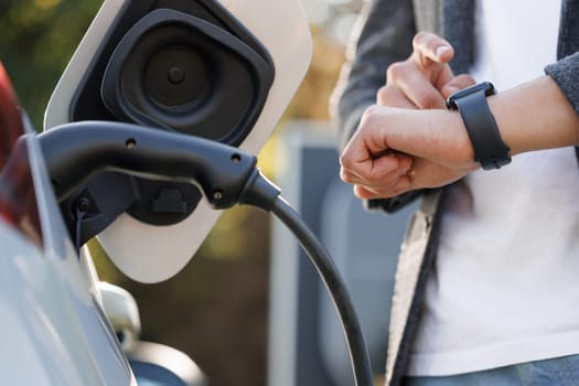 Businessman plugging in charging cable to to electric vehicle. Male hand inserts power connector into EV car and charges batteries, uses smartwatch for activates start charging.