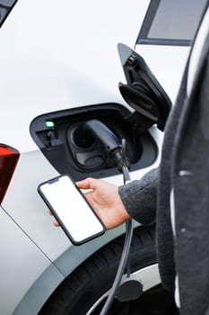 Businessman traveling by electric car having stop at chraging station standing holding smartphone looking at screen copy space for text or product close-up while having vehicle fully charged