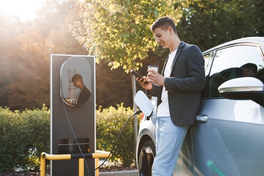 Caucasian businessman using smart phone and waiting power supply connect to electric vehicles for charging the battery in car. Plug charging an Electric car from charging station. Charging technology.