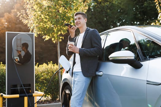 European man using smartphone voice messages and waiting power supply connect to electric vehicles for charging the battery in car. Plug charging an electric car from charging station.