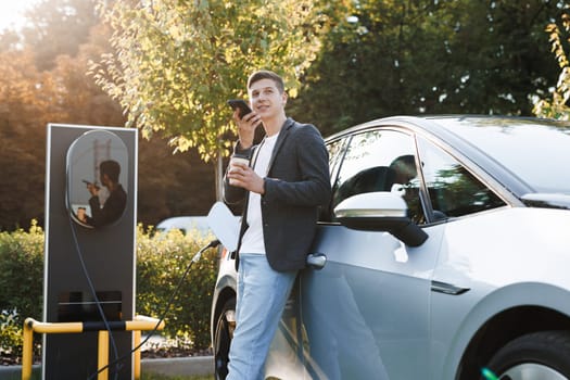 Businessman using smartphone voice messages and waiting power supply connect to electric vehicles for charging the battery in car. Plug charging an electric car from charging station.