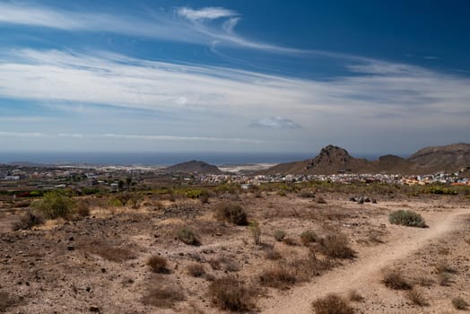 Hiking trail in a desert with a view of a seaside city, sea or ocean on a sunny day. Little mountains or hills at the horizon, blue sky and clouds. Little shrubs and a walking path between