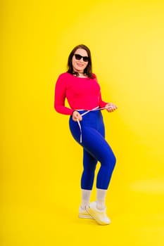 Pretty female with excess weight in a sporty top measuring waist over white yellow background