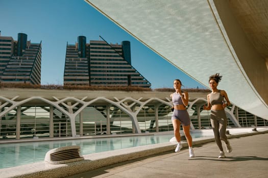 Two active women athlete running side by side along an outdoor track on modern buildings background