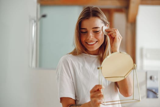 Smiling woman wiping her face with cotton pad in the bathroom. Skin care concept