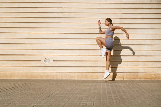 Female athlete in sportswear leaping in air with wall backdrop outdoors. Healthy lifestyle concept