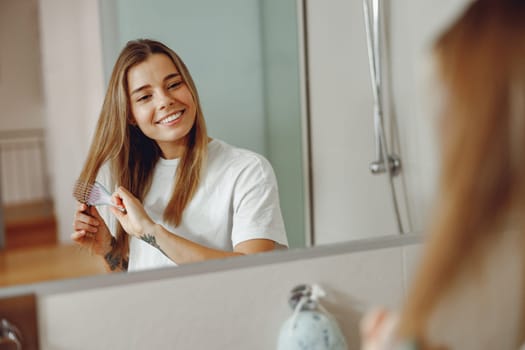 Woman standing in front of mirror at bathroom and combing her hair with brush after shower