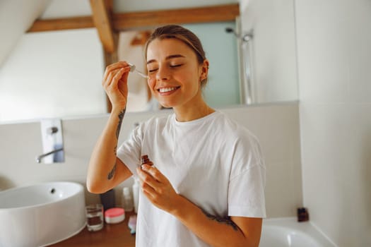 Beautiful young woman applying cosmetic serum onto her face in bathroom. Facial Skincare