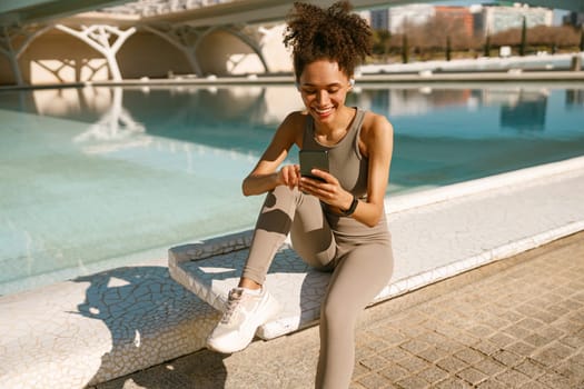 A young woman in sportswear is resting after a workout outside while sitting with smartphone