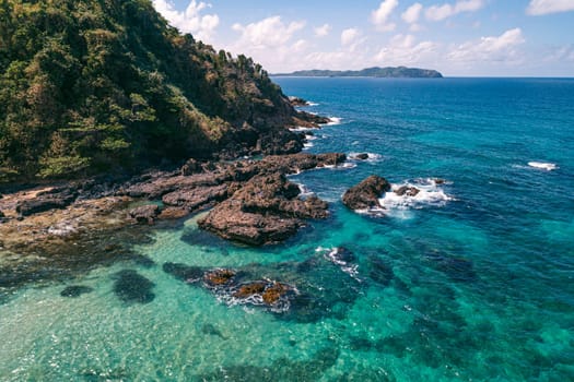 Aerial view of waves break on dark rocks near beach. Ocean waves crashing against rocky shore.