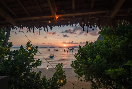 A serene view of a sunset from beneath the shelter of a beachside cafe, with fishing boats dotting the calm sea and silhouettes of distant islands on the horizon.