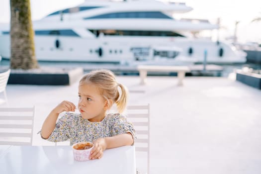 Little girl eats ice cream with a spoon, holding a glass with her hand on a table on the seashore. High quality photo