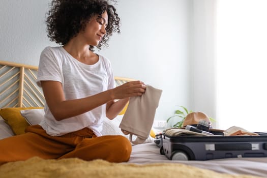 Young African American woman sitting on bed packing suitcase for summer holidays abroad. Copy space. Holiday, tourist, vacation concept.