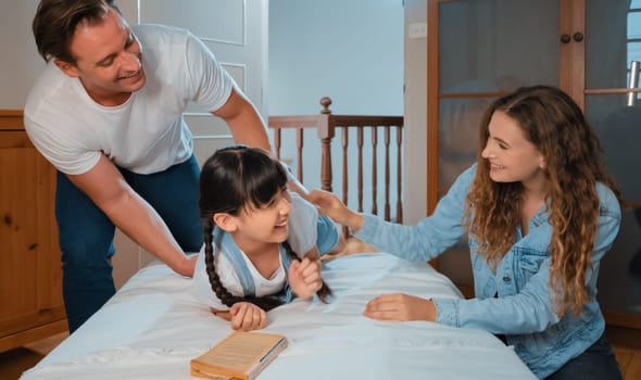 Happy modern family wakes up their little girl on the weekend with playful tickle expressing their love and affection for their young daughter, laughing and smiling together. Synchronos