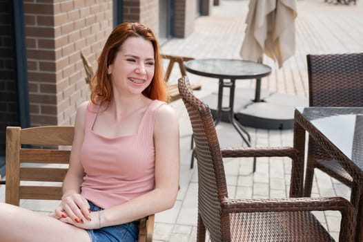 Beautiful young red-haired woman with braces on her teeth smiling while sitting in an outdoor cafe