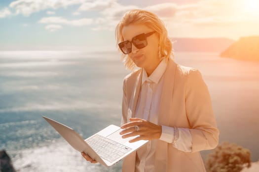 Freelance women sea. She is working on the computer. Good looking middle aged woman typing on a laptop keyboard outdoors with a beautiful sea view. The concept of remote work