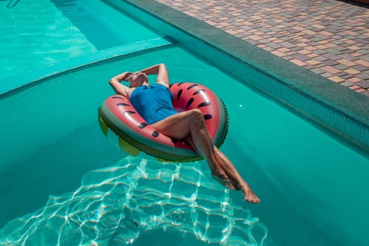 Happy woman in a swimsuit and sunglasses floating on an inflatable ring in the form of a watermelon, in the pool during summer holidays and vacations. Summer concept