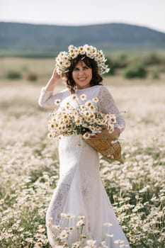 Happy woman in a field of daisies with a wreath of wildflowers on her head. woman in a white dress in a field of white flowers. Charming woman with a bouquet of daisies, tender summer photo.