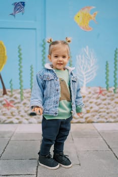 Little smiling girl stands near colorful painted wall. High quality photo