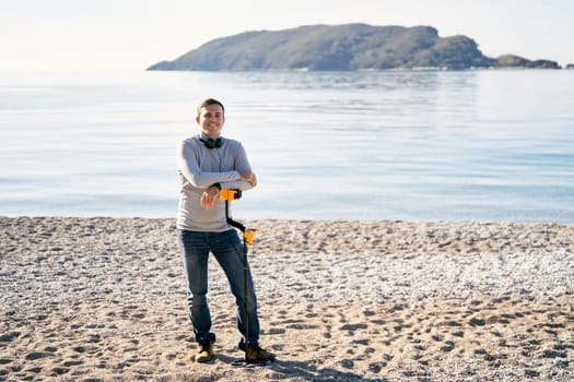 Smiling man stands leaning on a metal detector on the shore. High quality photo